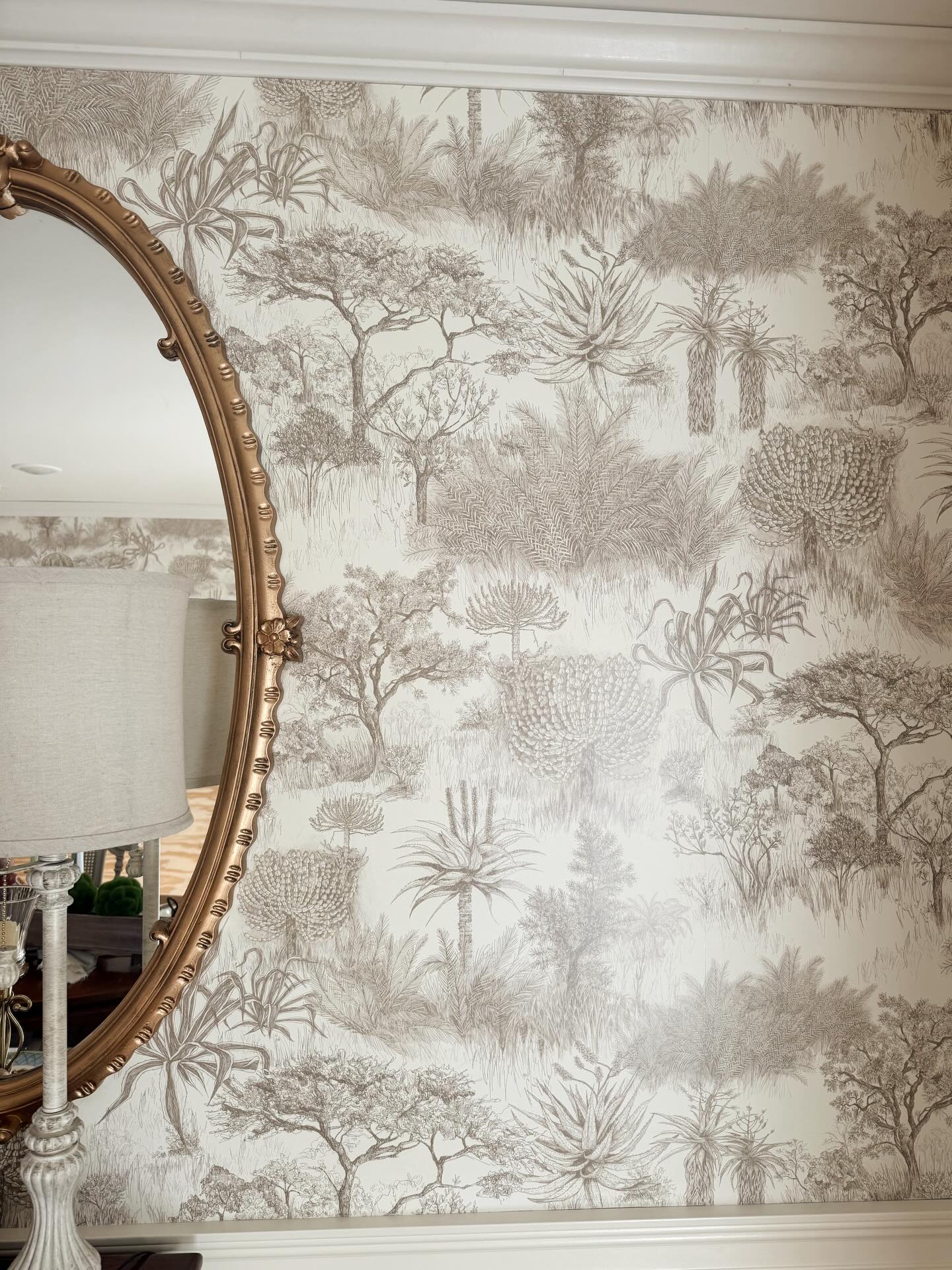 A gold ornate mirror on a wall covered in beige wallpaper with a brown, hand-drawn botanical pattern of trees and plants.