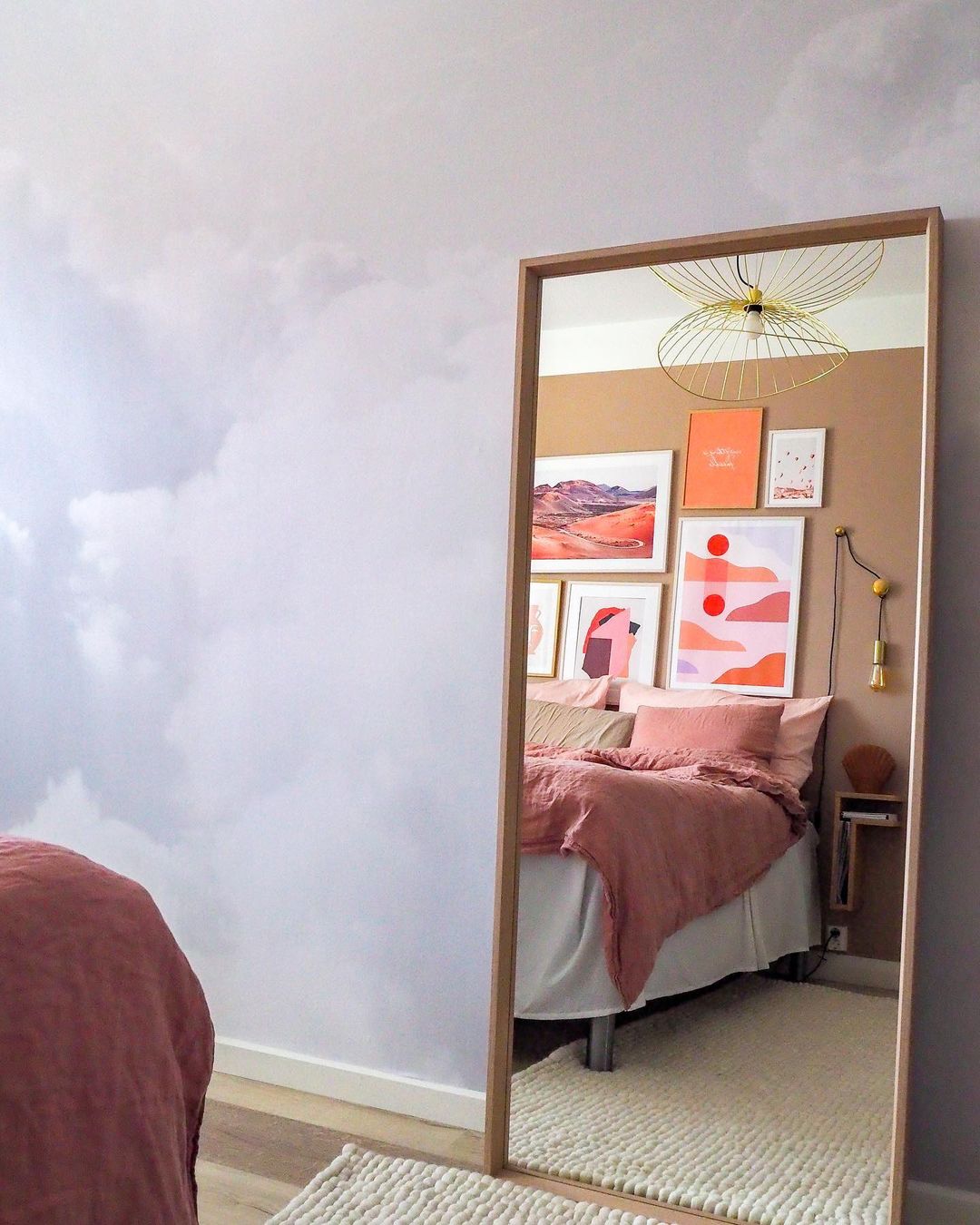 Bedroom interior reflected in a full-length mirror. Bed with pink linens, gallery wall, cloud wallpaper.