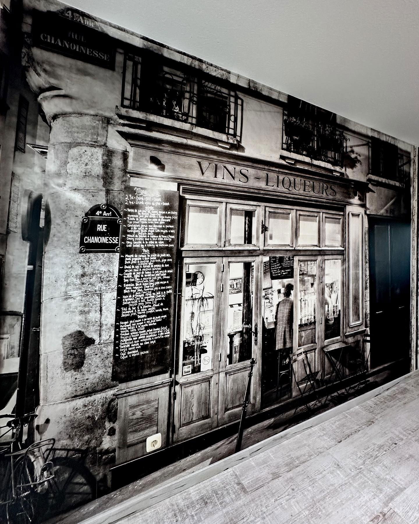 Zwart-wit muurschildering van een Parijse wijnwinkel, "Vins-Liqueurs," met een menubord en straatnaambord "Rue Chanoinesse".