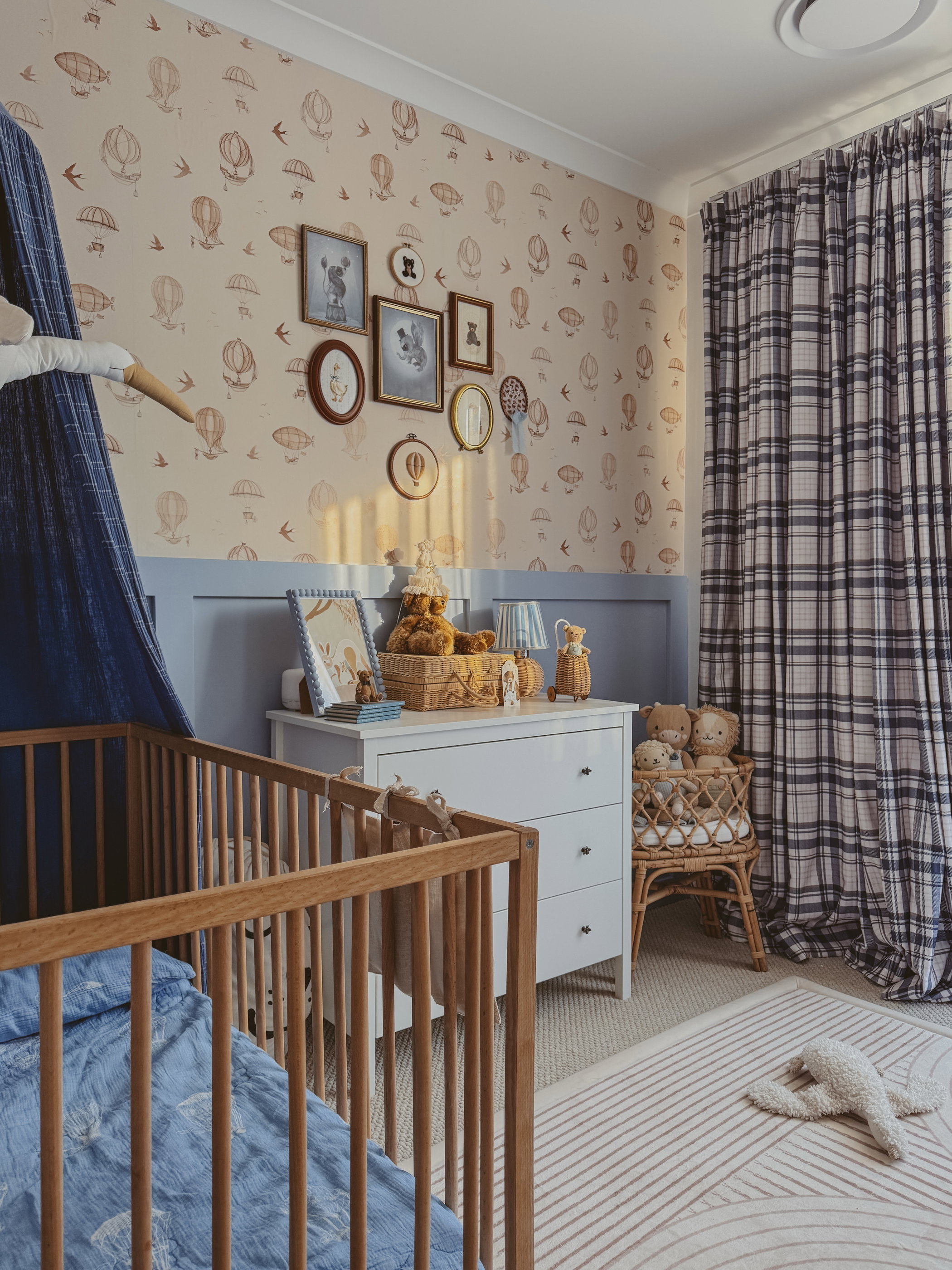 Chambre de bébé avec berceau, commode, ours en peluche, papier peint montgolfière et rideaux à carreaux.