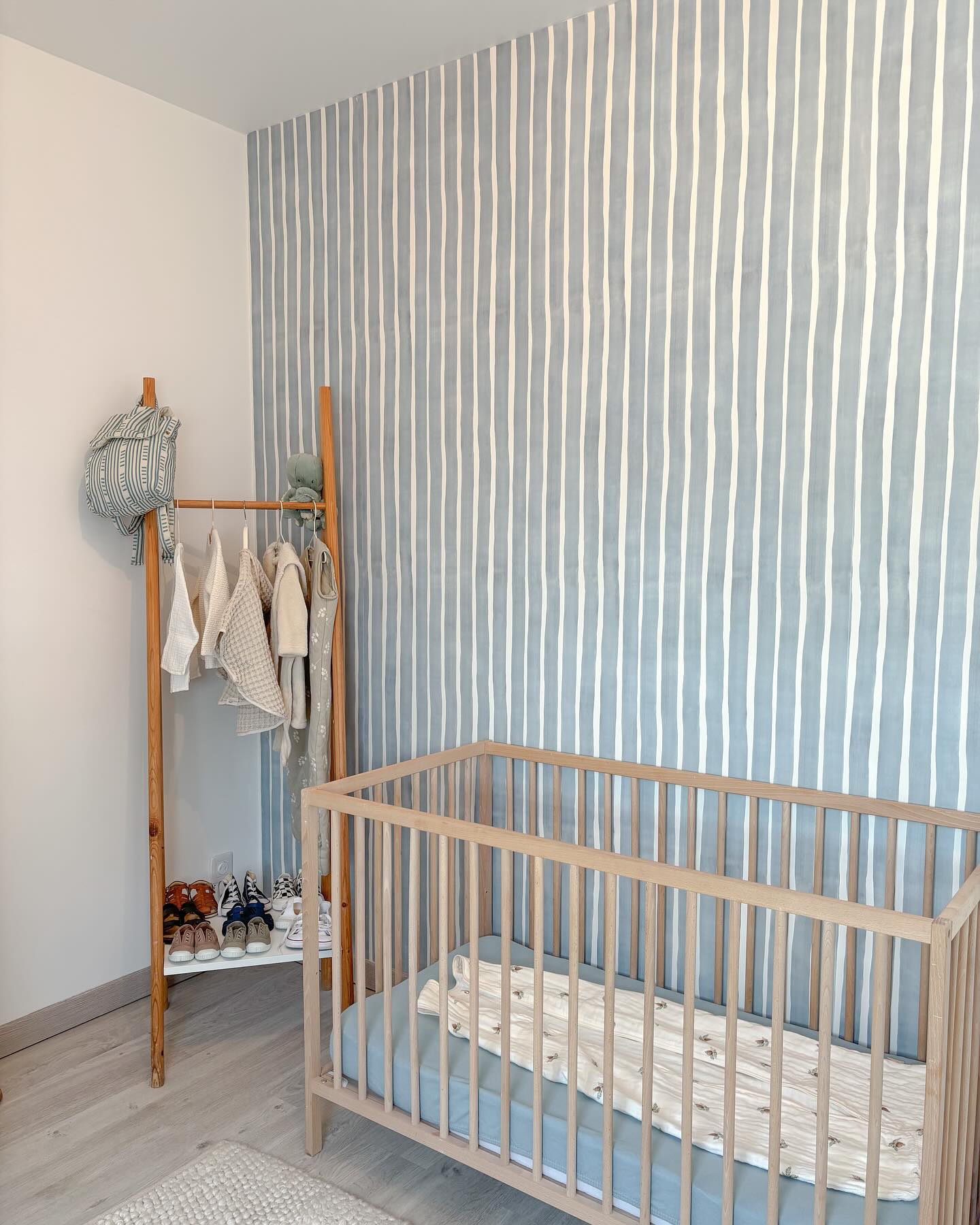 Nursery with wooden crib, blue striped wallpaper, and clothes rack with baby clothes and shoes.