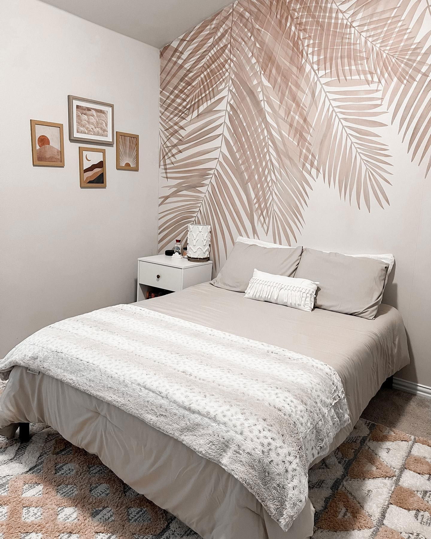 Dormitorio con papel tapiz de hojas de palma, ropa de cama neutra y arte enmarcado sobre una mesita de noche blanca.