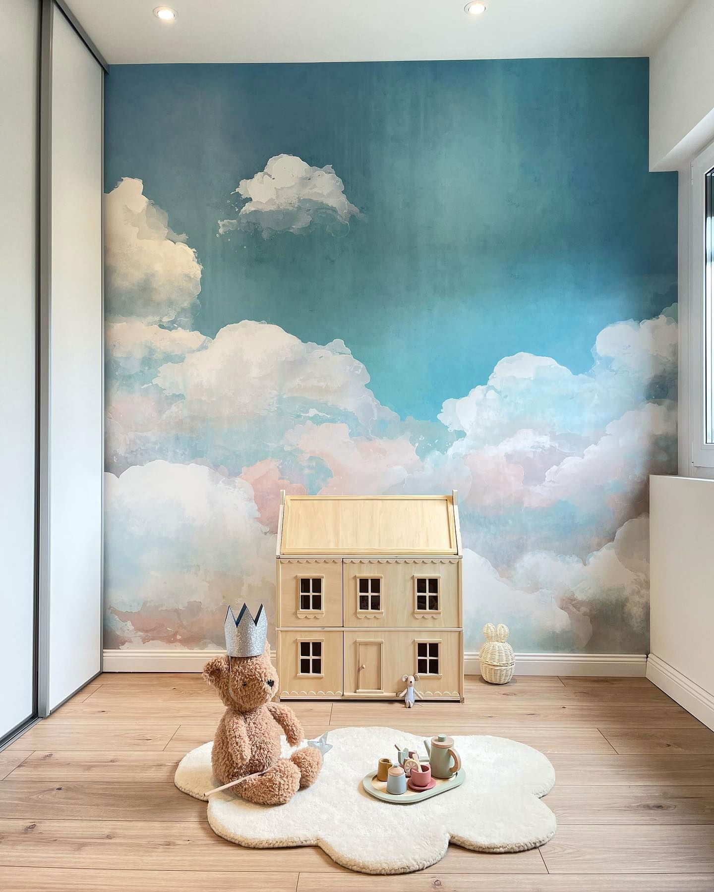 Child's room with cloud wallpaper, wooden dollhouse, and teddy bear wearing a crown on a flower-shaped rug.