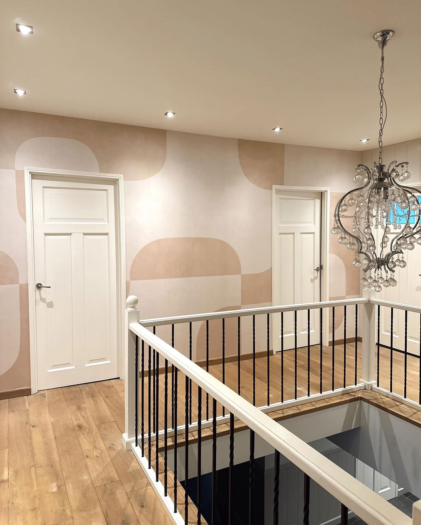 Flur mit Holzböden, weißen Türen und einer geometrisch gemusterten Wand. Ein Kristalllüster hängt von der Decke.