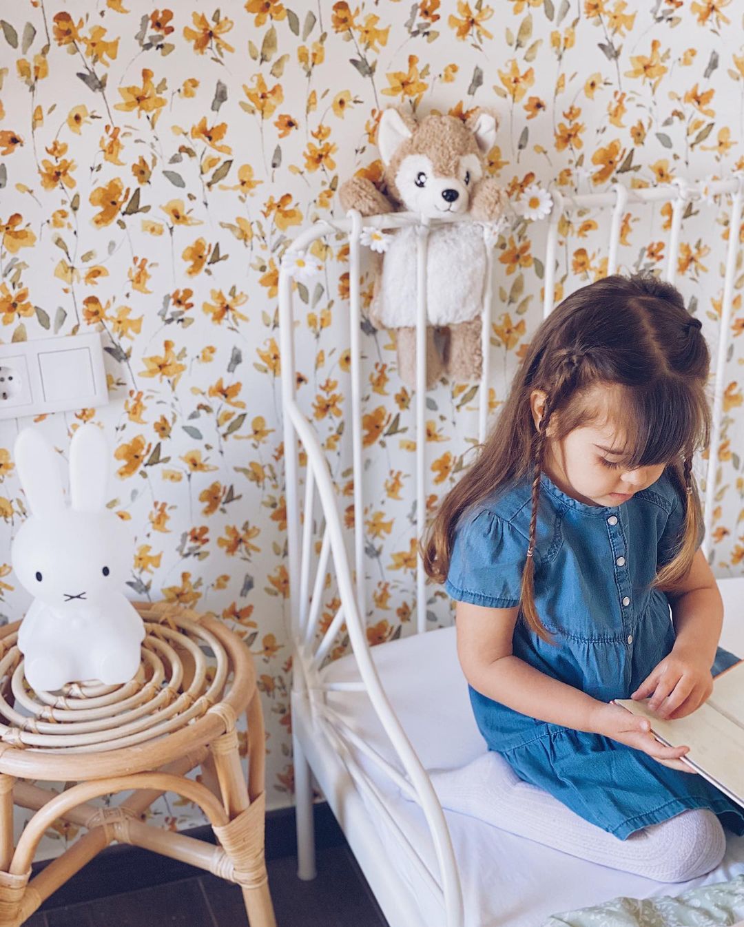 A young girl in a blue dress sits on a white bed reading a book. Floral wallpaper and a Miffy lamp are visible.