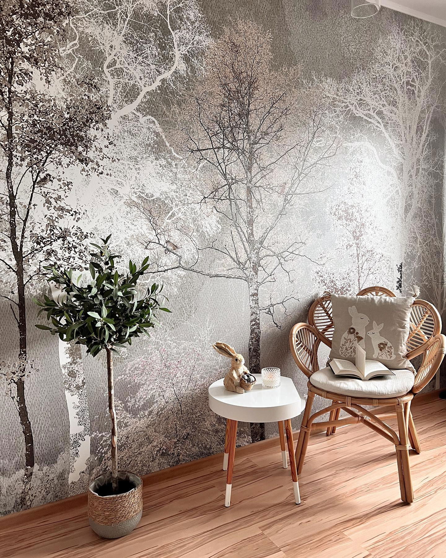 Cozy reading nook with forest wallpaper, rattan chair with bunny pillow, and small white table.