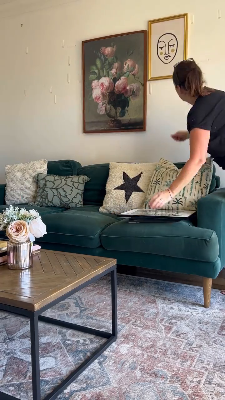 Living room scene with a woman arranging art above a green velvet couch.
