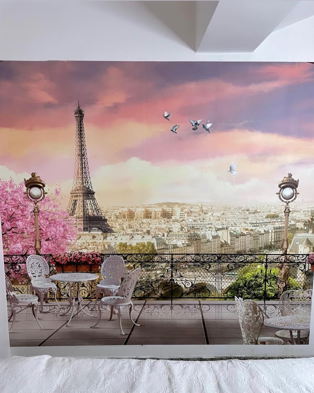Parijse muurschildering met de Eiffeltoren, een witte bistroset en een smeedijzeren balkon tegen een roze en paarse lucht.