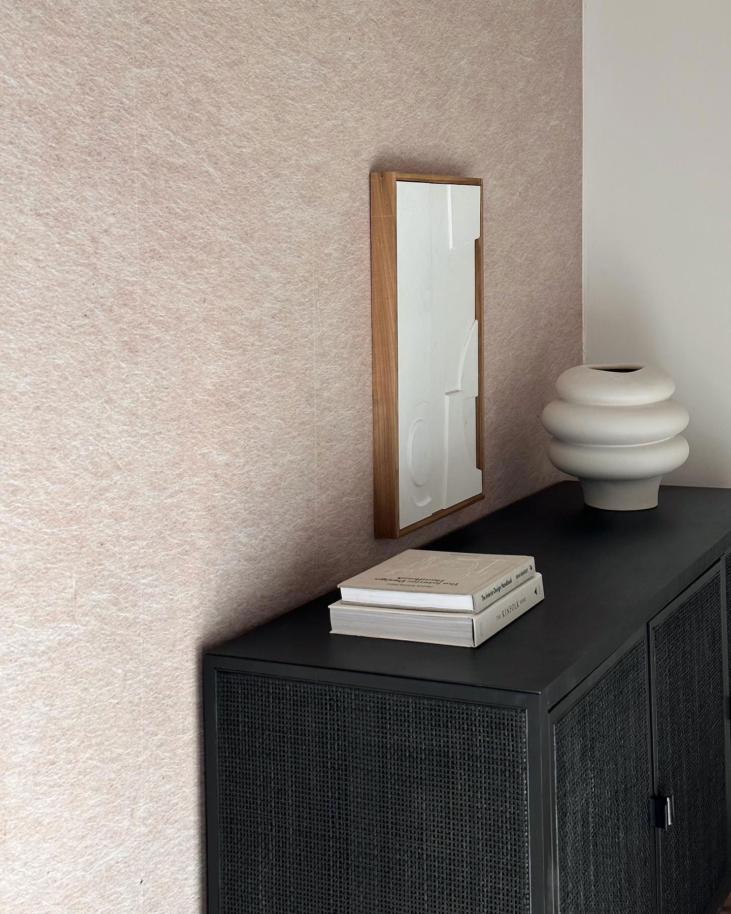 Intérieur avec une armoire noire, des livres, un vase et une œuvre d'art encadrée sur un mur rose texturé.