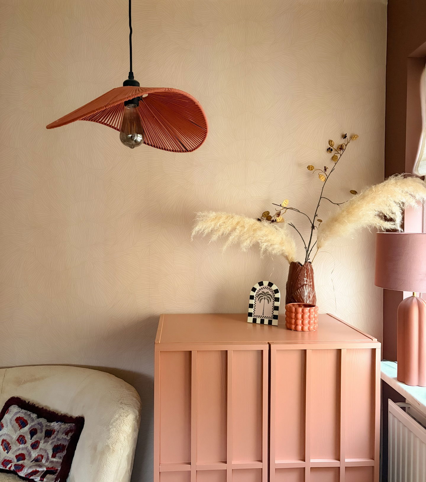 Coral pendant light hangs above a coral cabinet with decorative objects and pampas grass.