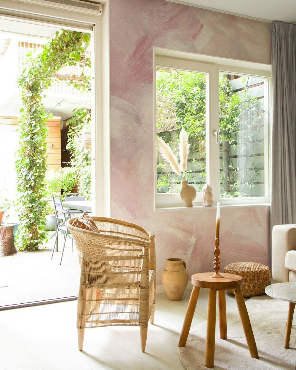 Living room with pink brushstroke wall, rattan chair, and garden view through open door.