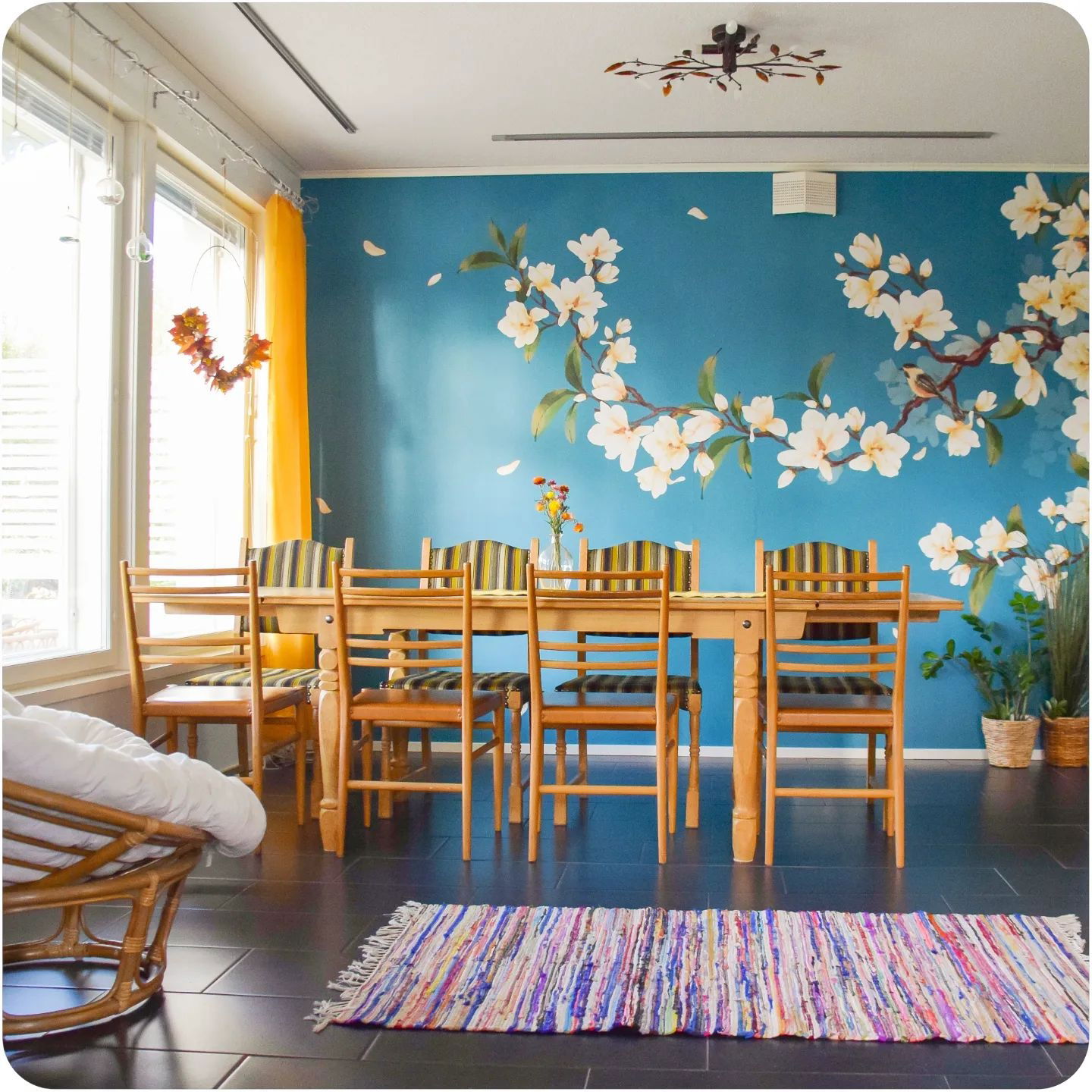 Dining room with blue floral mural, wooden table and chairs, and colorful rug.