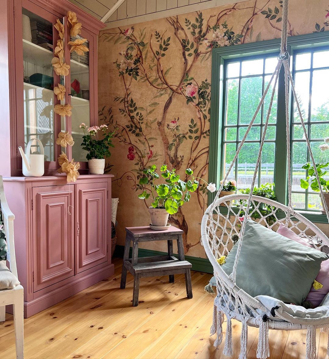 Hyggeligt værelse med blomstret tapet, pink skab og en hvid macrame-gyngestol med puder.