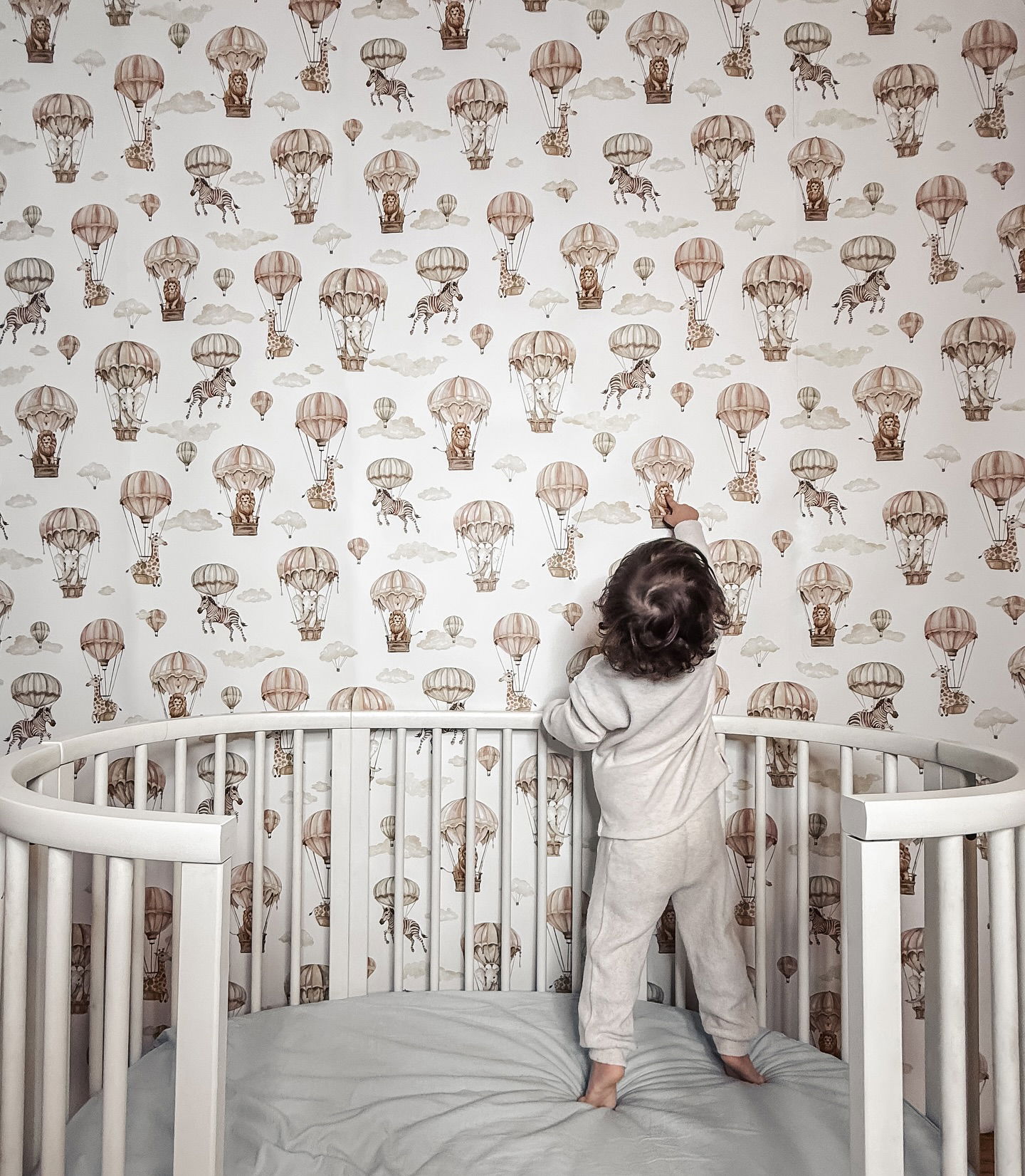 Child in crib reaching for wallpaper. The wallpaper has a pattern of hot air balloons and animals.