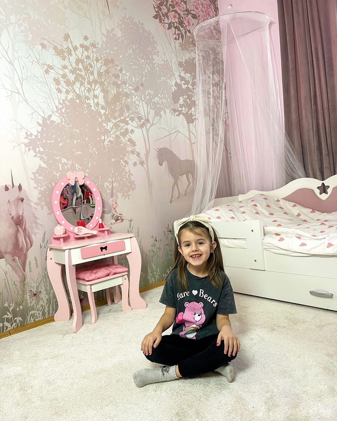 A young girl sits cross-legged in a pink-themed bedroom with a Care Bears t-shirt and headband.
