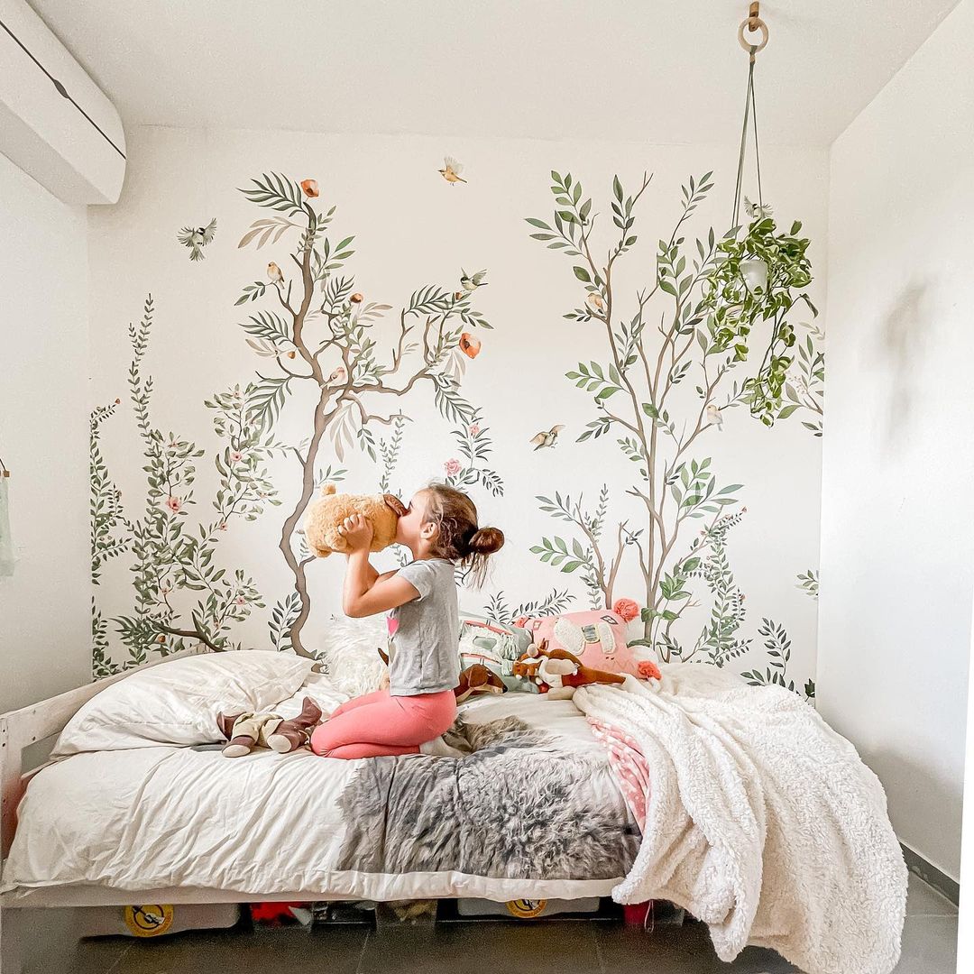 A girl hugs a stuffed animal on a bed with a floral mural. The room has a cozy, nature-inspired aesthetic.