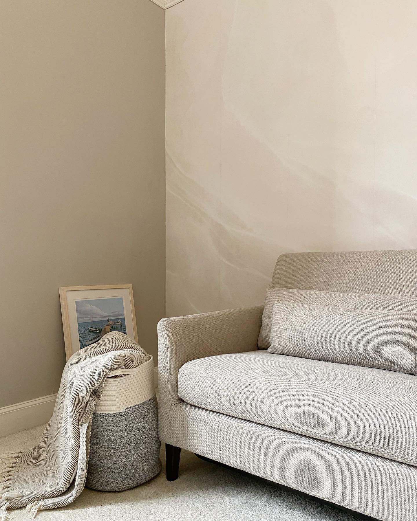 Angolo neutro del soggiorno con divano beige, cesto intrecciato con plaid e arte incorniciata.
