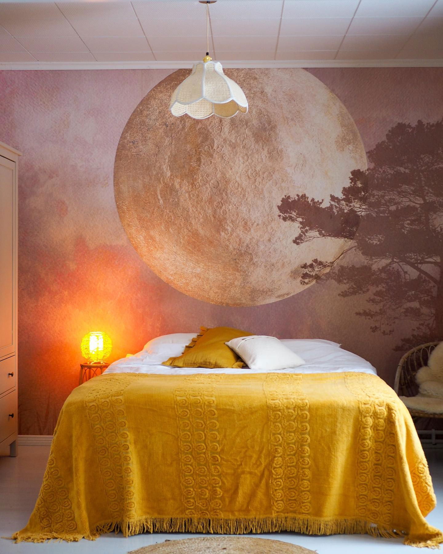 Schlafzimmer mit gelber Decke, weißen Laken und Mondbild. Über dem Bett hängt eine gewebte Pendelleuchte.