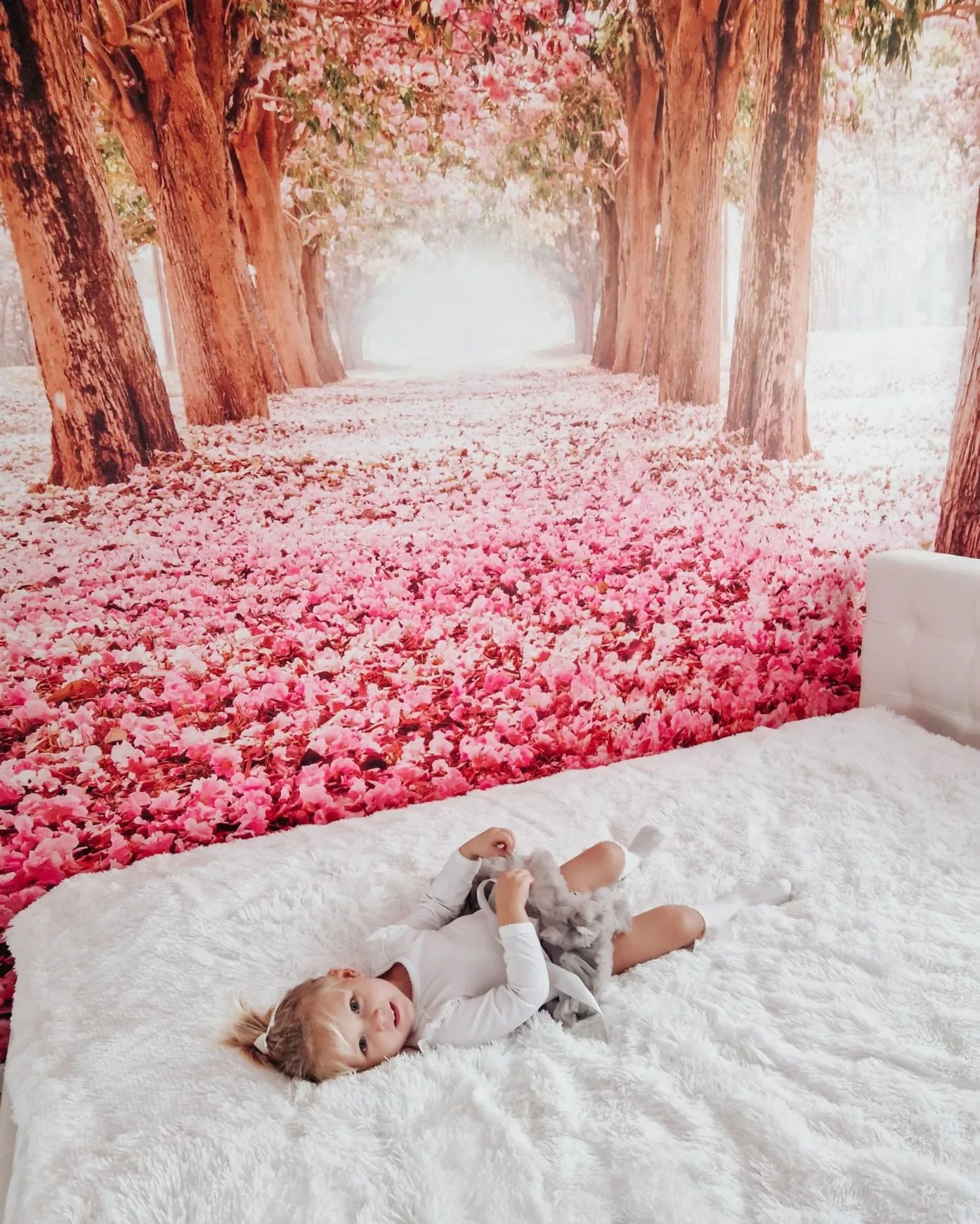 A child lies on a white bed with a floral backdrop. The girl is holding a gray stuffed animal.