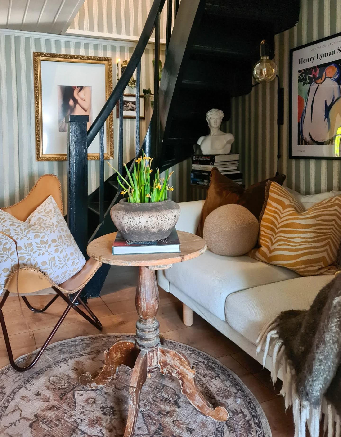 Living room with patterned wallpaper, a staircase, sofa, accent chair, and antique table with flowers.