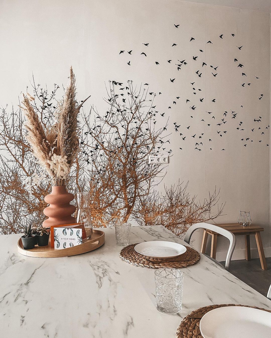 Marble dining table set with vase of pampas grass, plates, and glasses. Wall art of trees and birds in background.