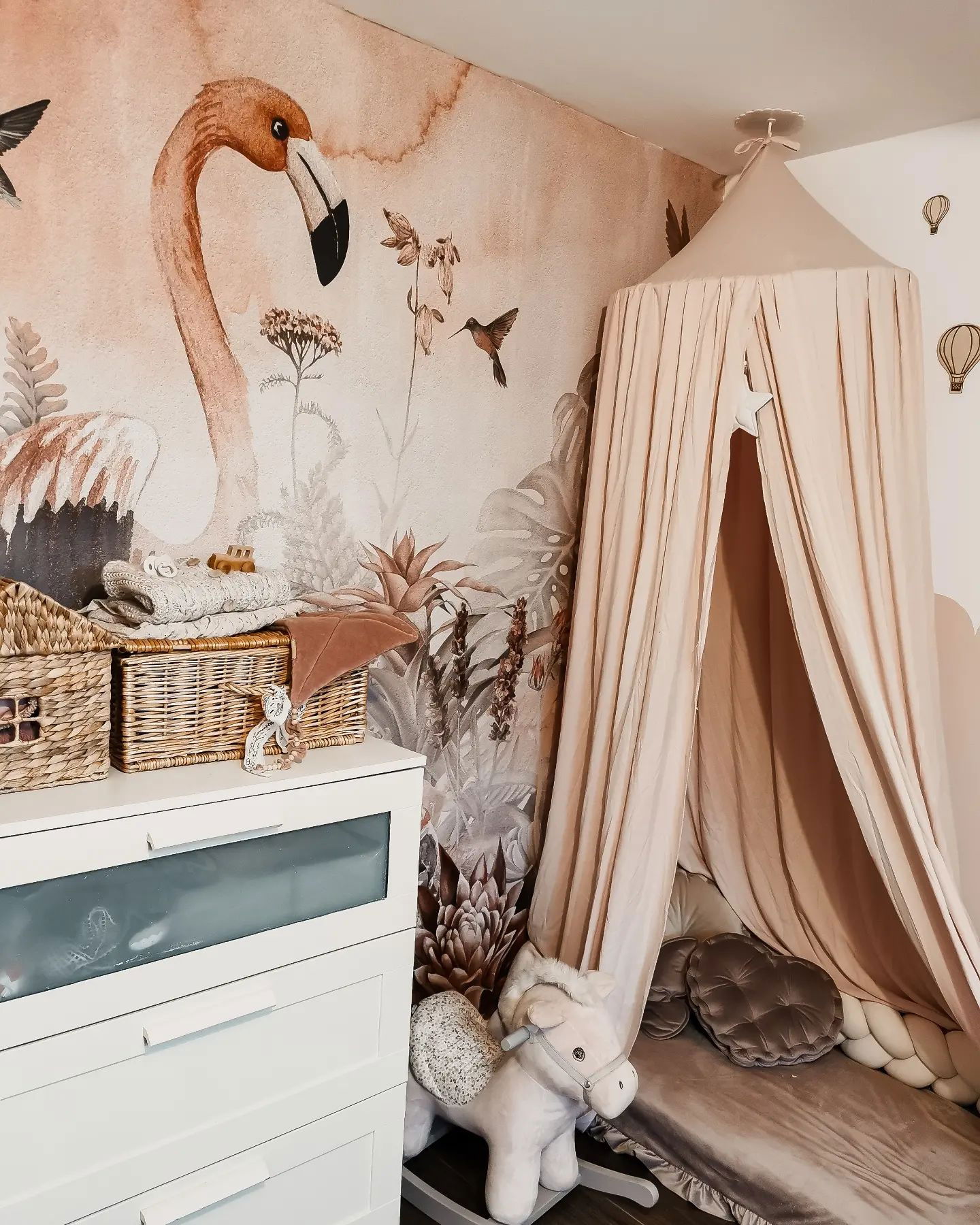 Nursery with flamingo wallpaper, white dresser with wicker baskets, and a beige canopy bed with pillows.