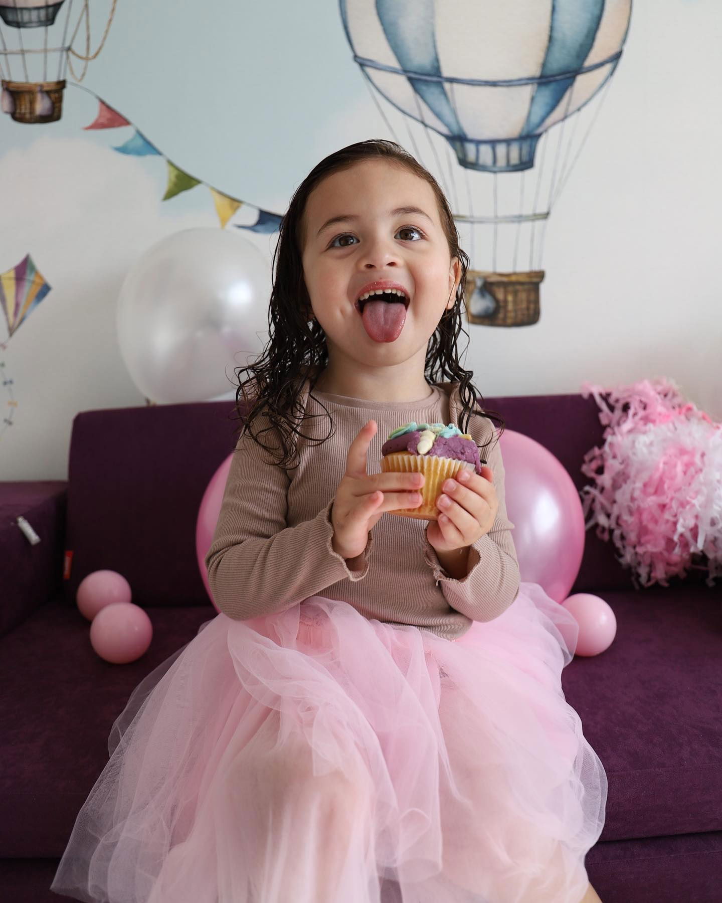 Une jeune fille en tutu rose est assise sur un canapé violet, tenant un cupcake et tirant la langue. Des ballons et des décorations l'entourent.