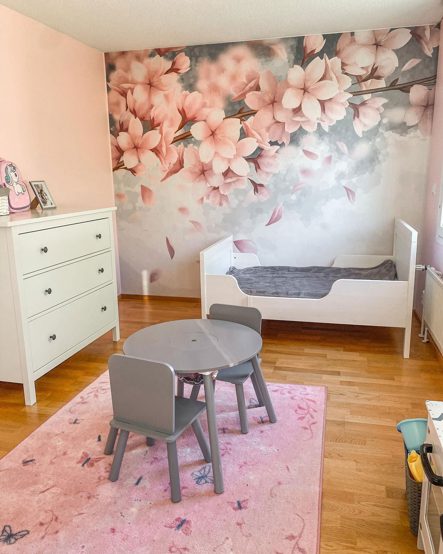 Child's bedroom with pink floral wallpaper, white bed, dresser, and gray table with chairs on a pink butterfly rug.