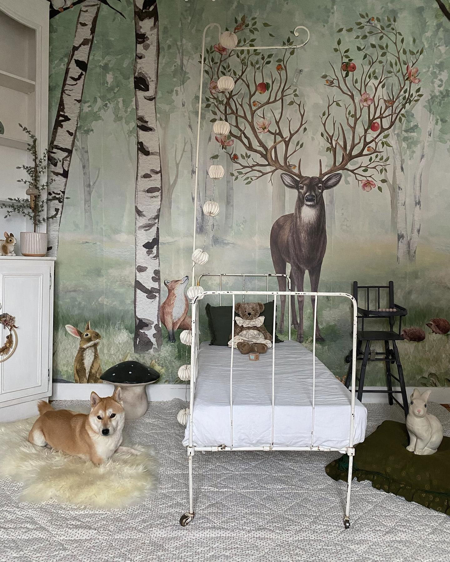 Whimsical nursery with forest mural, white metal bed, and Shiba Inu on a sheepskin rug.