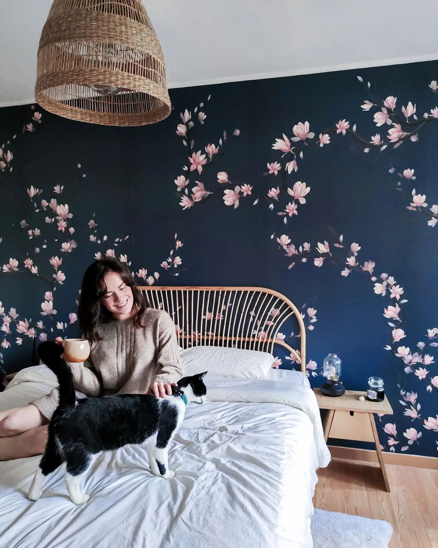 A woman sits in bed with a black and white cat, holding a mug in a bedroom with floral wallpaper.