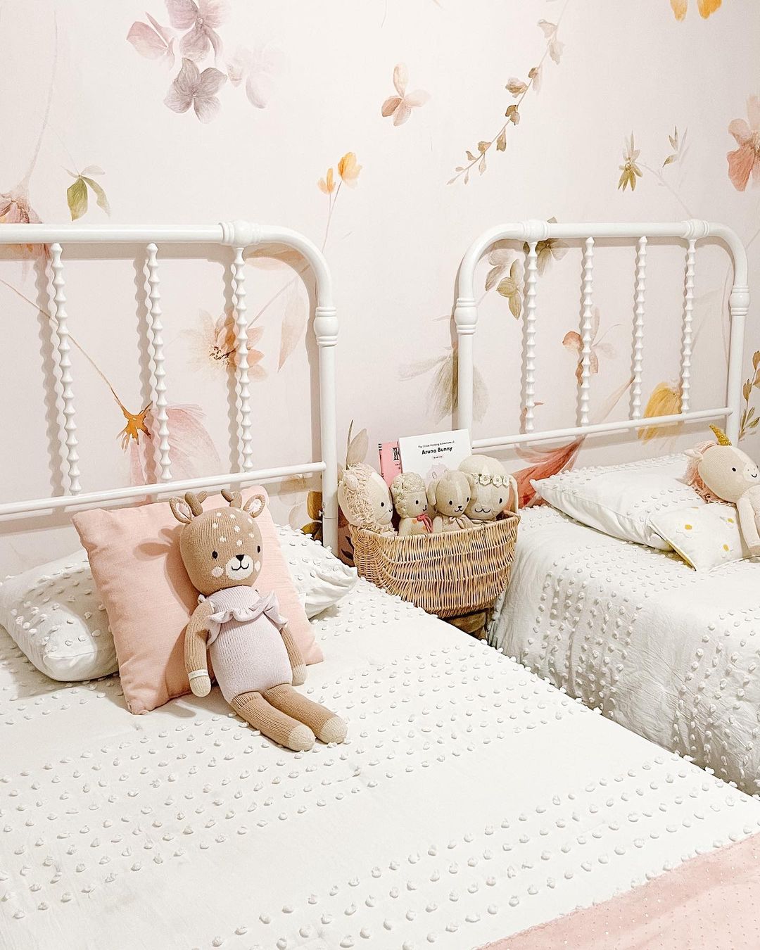 Dos camas blancas con papel tapiz floral, una cesta de animales de peluche y una muñeca de ciervo.