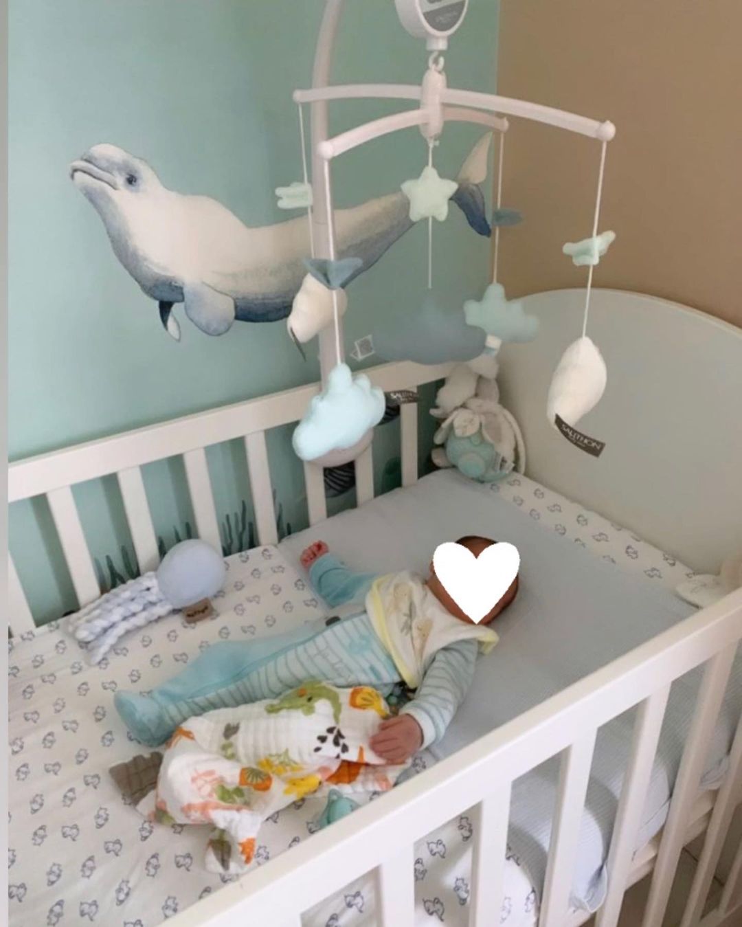 Baby in crib with mobile, blanket, and whale wall decal.