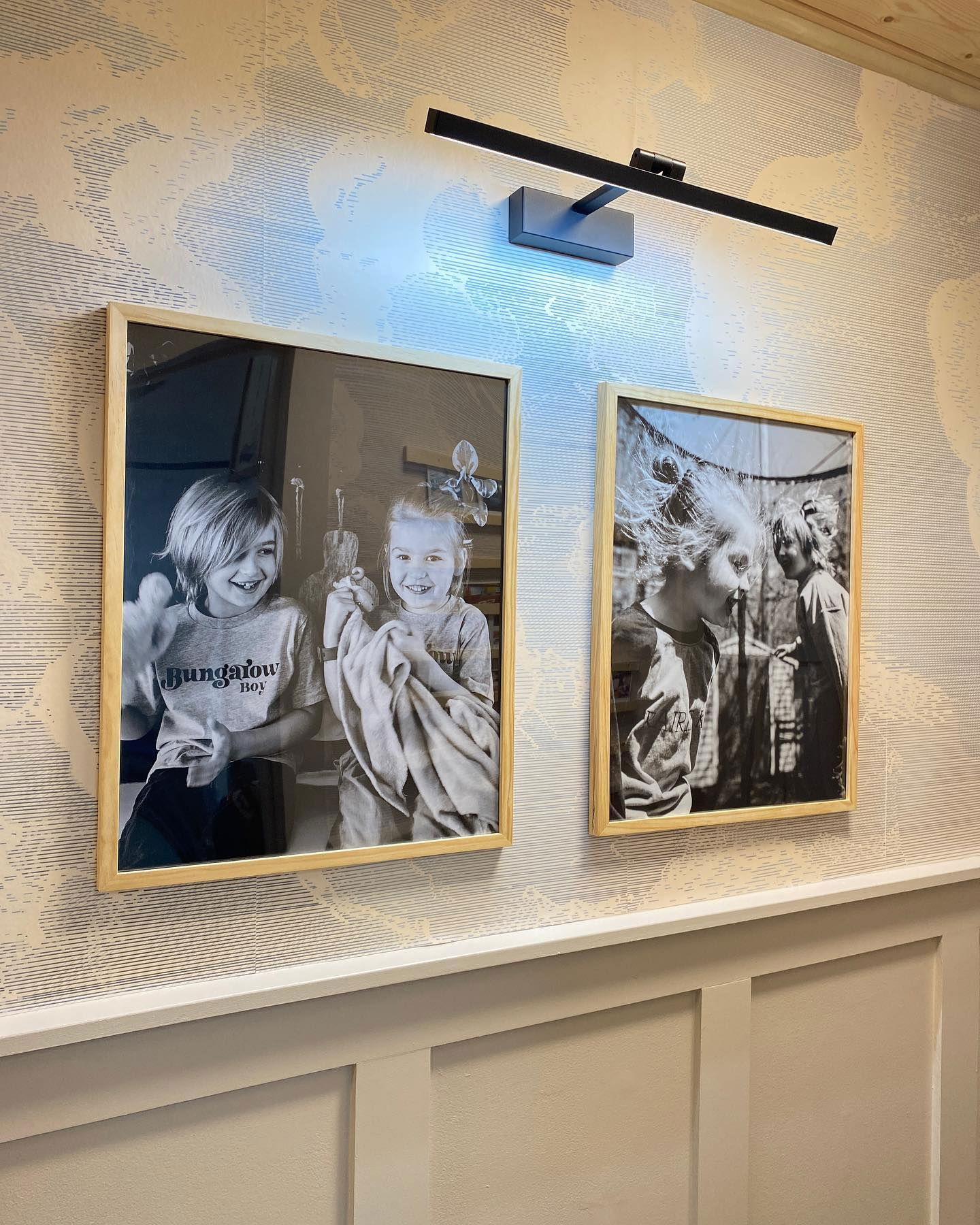 Zwei gerahmte Schwarz-Weiß-Fotos hängen an einer gemusterten Wand unter einer schwarzen Lampe.