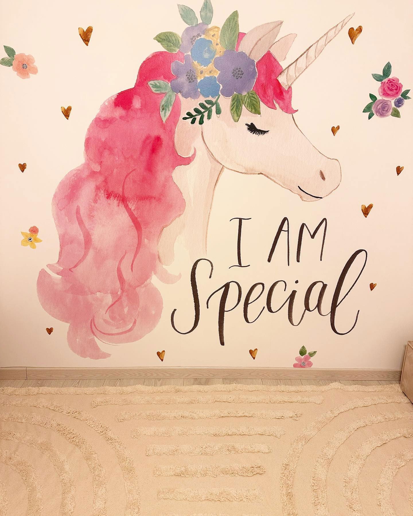 Aquarell-Einhorn-Wandbild mit rosa Mähne, Blumenkrone und dem Text "Ich bin besonders" an einer Wand über einem strukturierten Teppich.