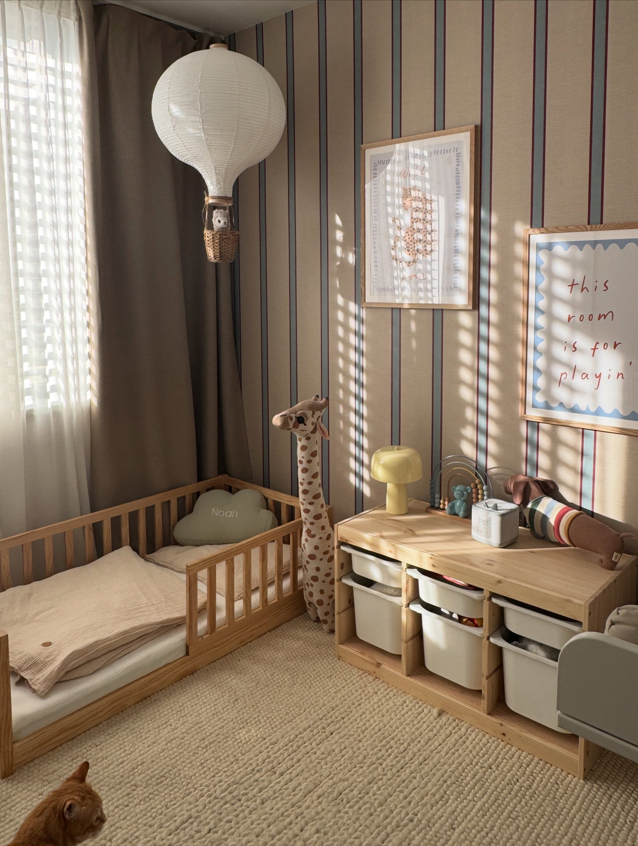 White hot air balloon lamp, wooden toddler bed, striped wallpaper, a wood storage unit, and a ginger cat.