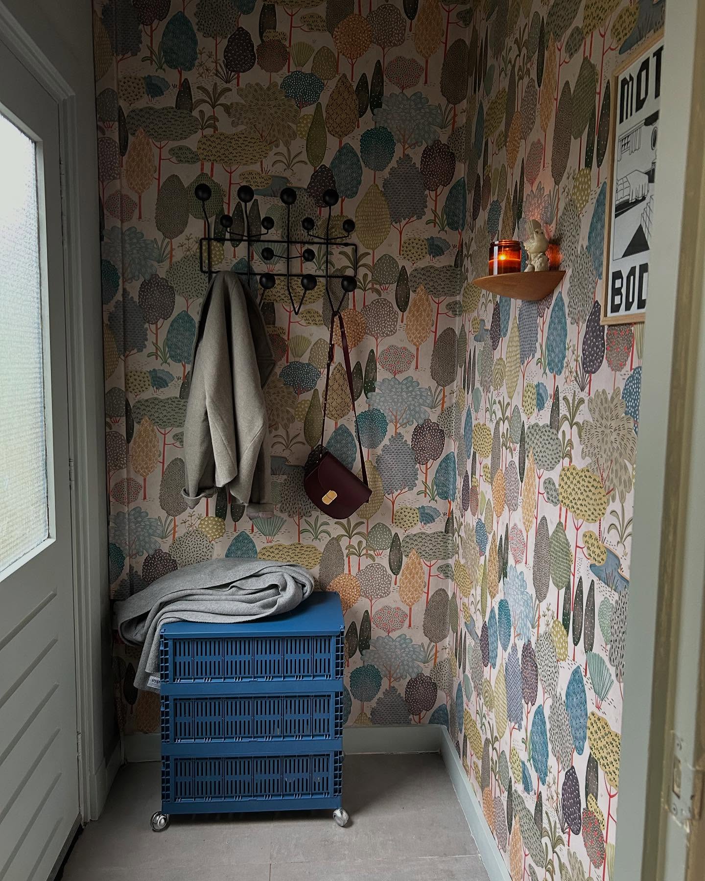 Hallway with colorful tree wallpaper, blue rolling storage, and coat rack with jacket and purse.