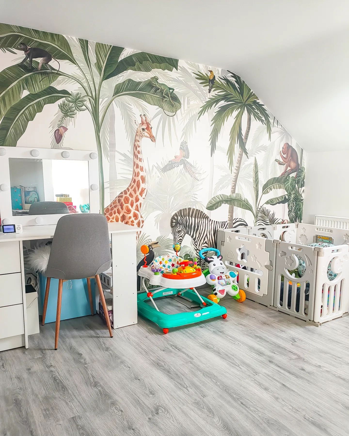 Nursery with jungle-themed wallpaper, white vanity with lighted mirror, gray chair, baby walker, and white playpen.