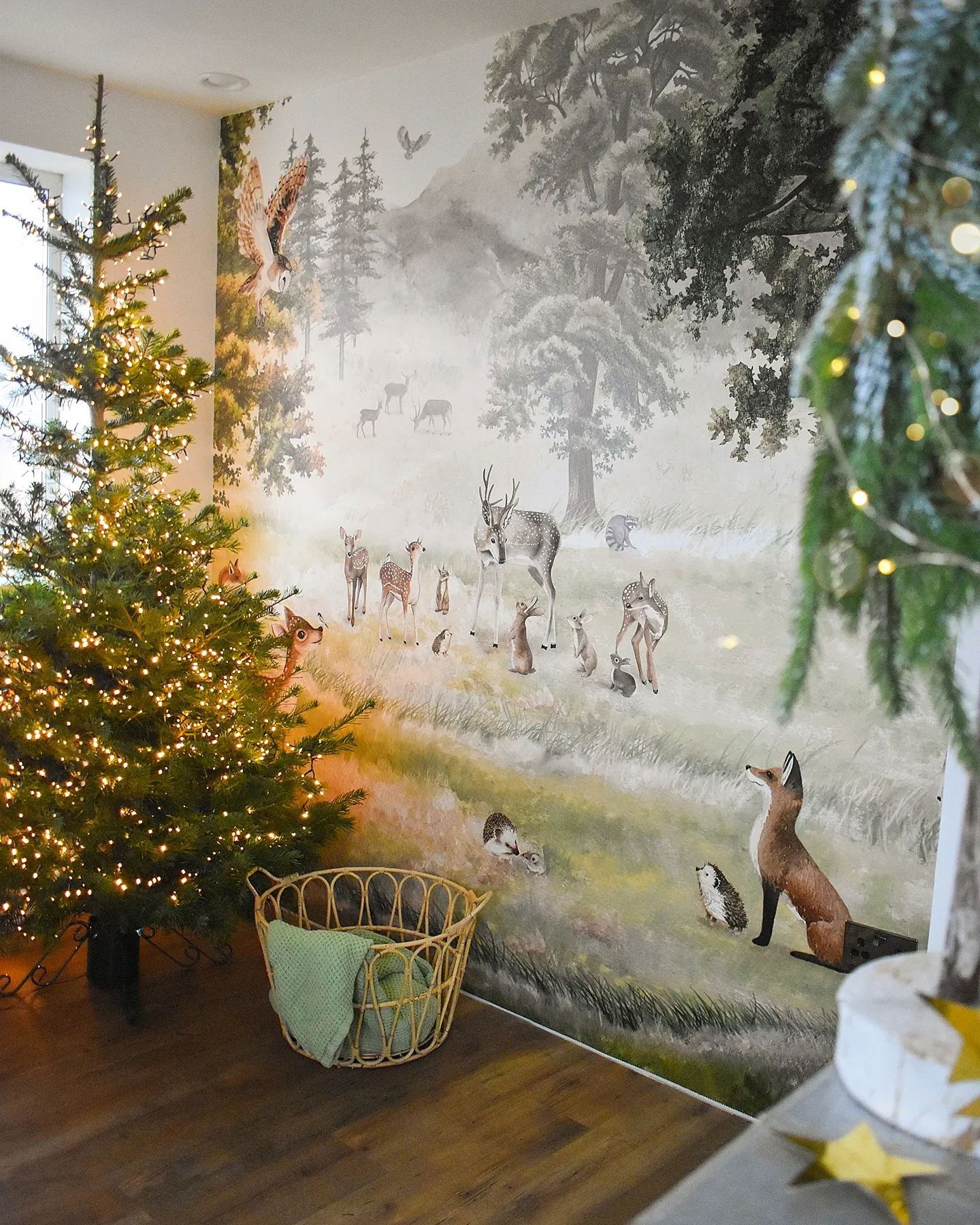 Pièce festive avec un arbre de Noël illuminé et une murale d'animaux des bois. Un panier en osier avec une couverture verte est posé près de l'arbre.