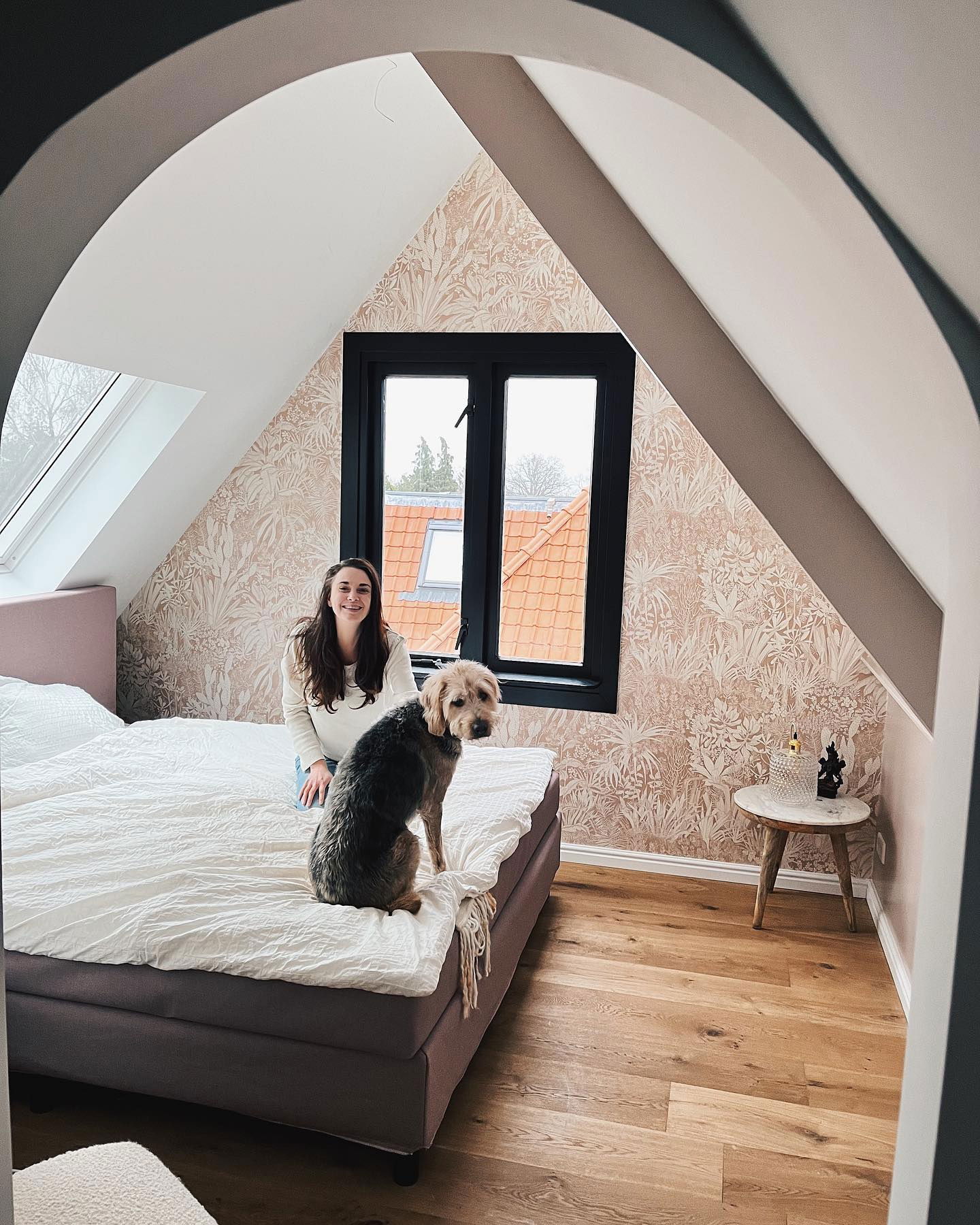 Una mujer y su perro están sentados en una cama en un dormitorio abuhardillado con papel tapiz floral.