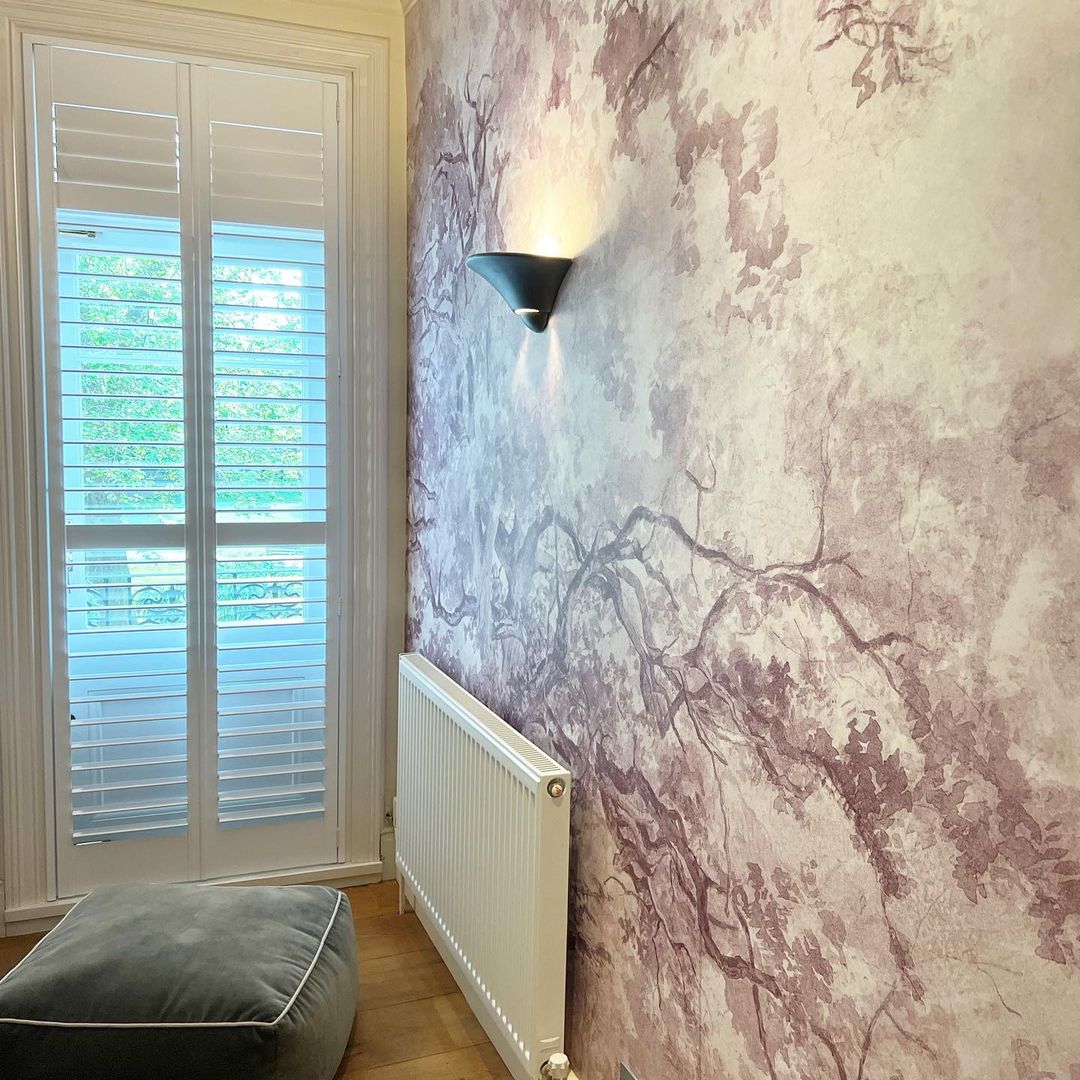 Coin intérieur avec papier peint à motif d'arbre, applique, radiateur et fenêtre à volets.