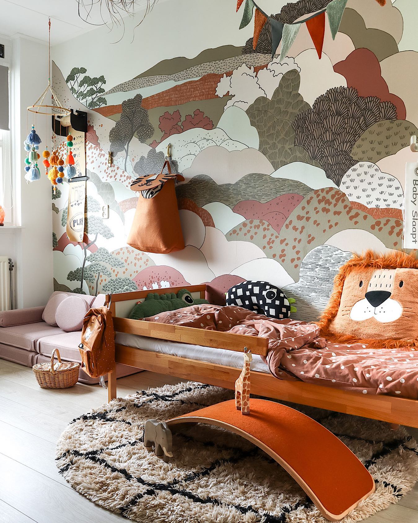 A child's bedroom featuring a wooden bed with a lion pillow. The wall has a mural of green, pink, and brown hills. A shaggy rug, an orange wobble board, and a pink floor mattress are visible.
