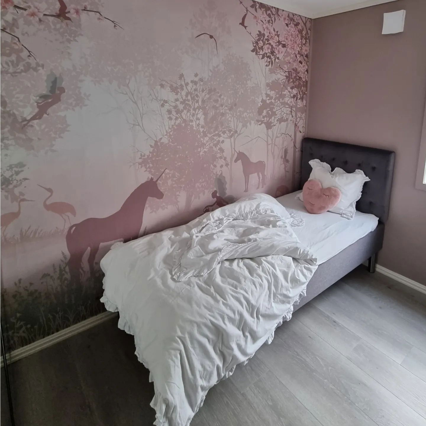 Bedroom with unicorn wallpaper, gray bed, white ruffled bedding, and a pink heart pillow.