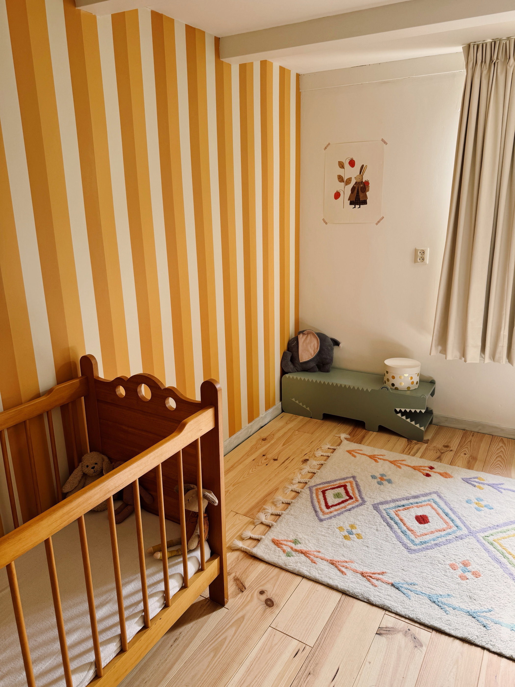 Lastenhuone, jossa on vaaleasta puusta tehty sänky, kelta-oranssiraitainen tapetti, vihreä krokotiilinmuotoinen penkki ja kuviollinen matto.