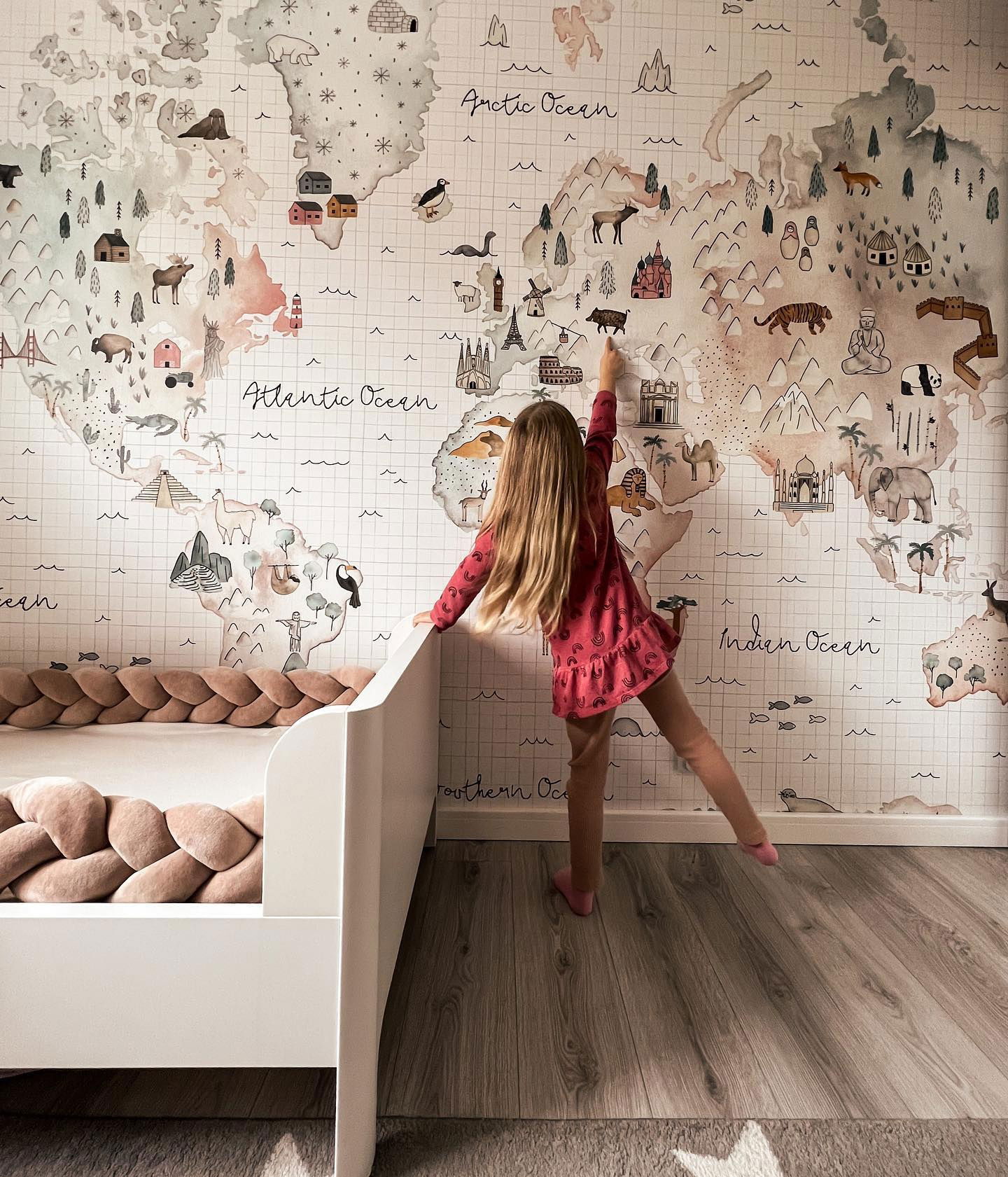 Child pointing at world map wallpaper in bedroom with white bed and braided bumper.