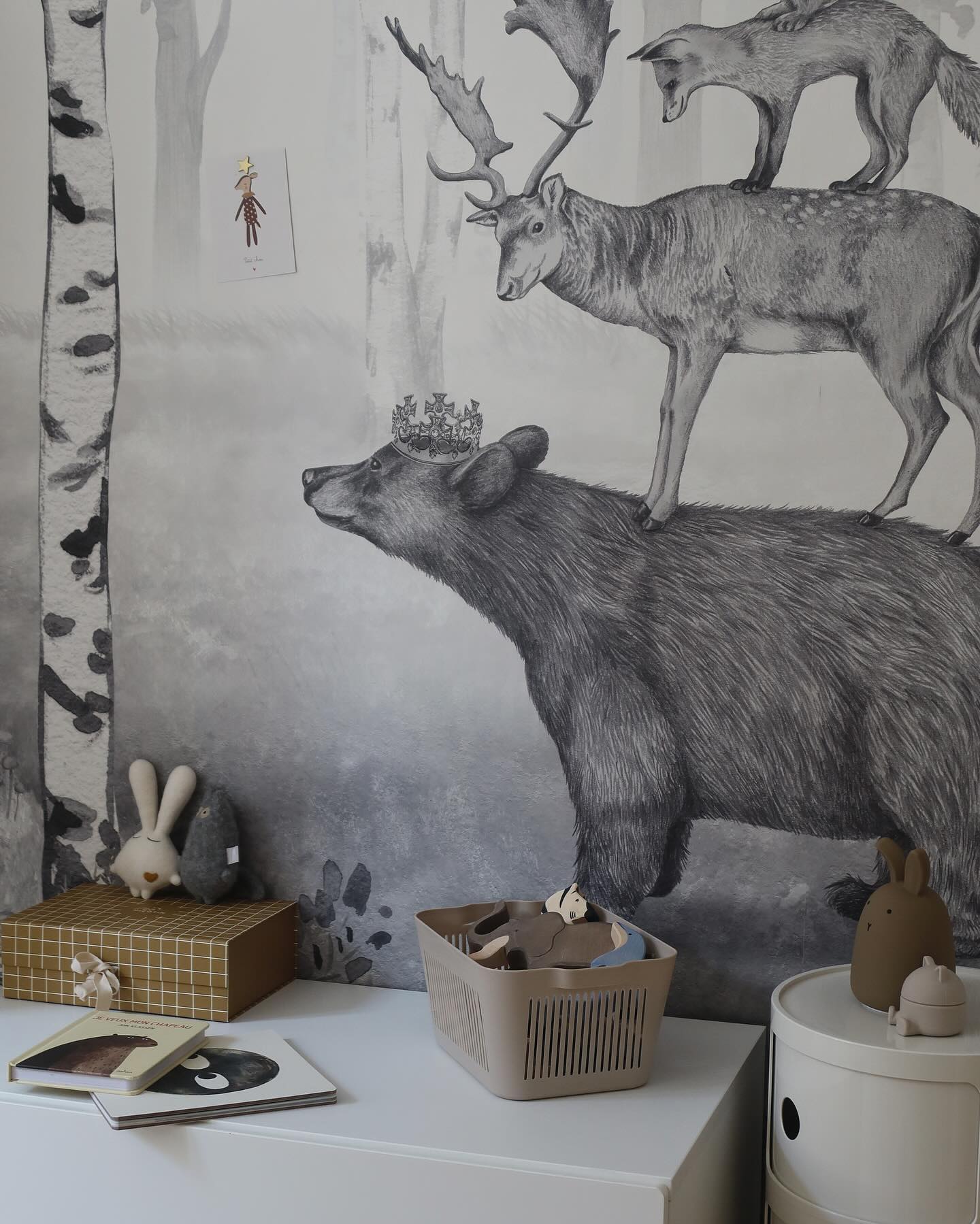 Child's room with animal-themed wallpaper, featuring a bear, deer, and fox. Toys and books are on a white dresser.