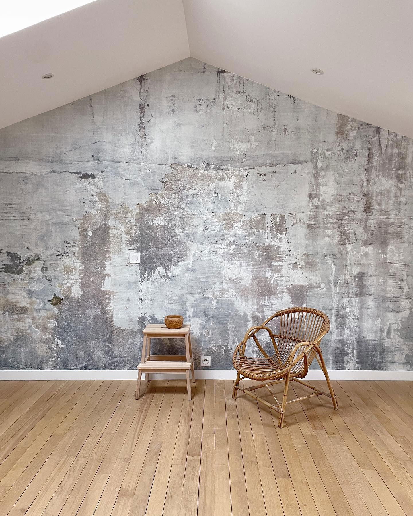 Minimalistische kamer met rotan stoel, houten kruk en grijze muur met textuur.
