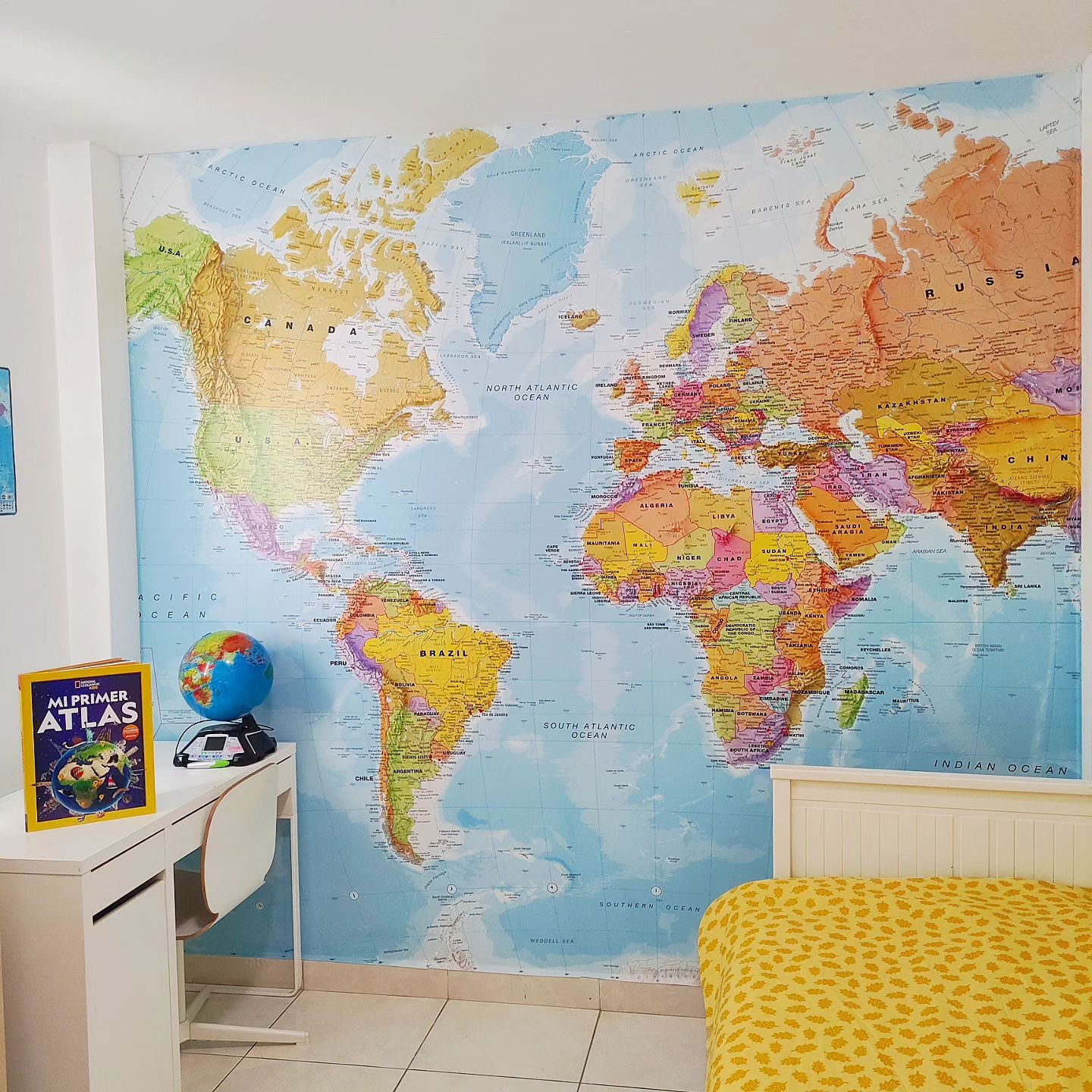 Child's room with world map wall mural, desk with atlas, and yellow bedspread.