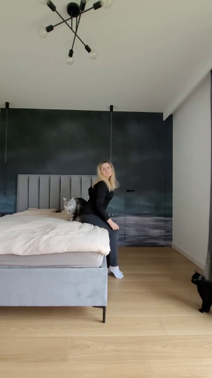 A woman sits on a gray bed with two cats in a modern bedroom.
