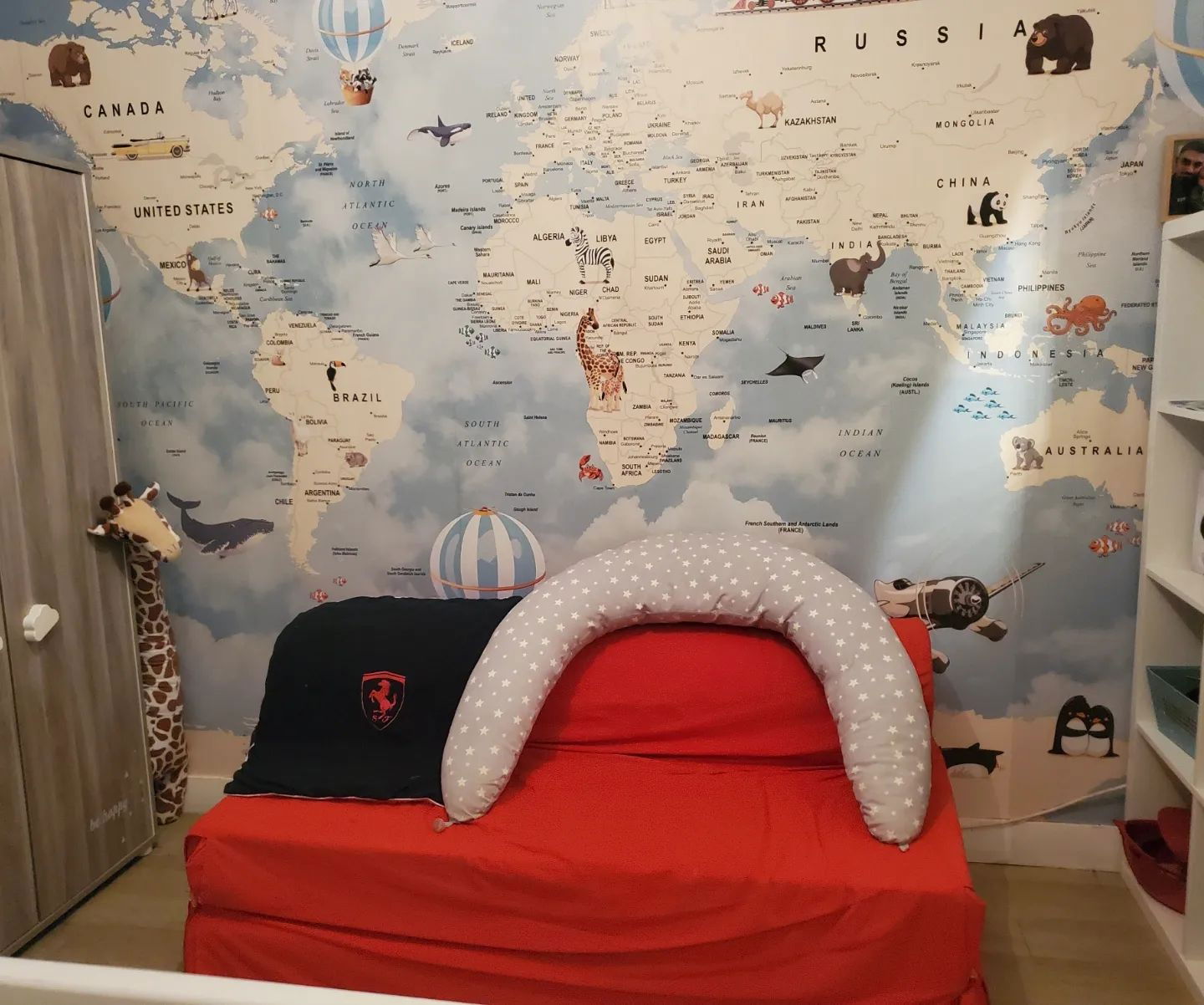 Stanza di bambino con carta da parati a tema mappamondo, divano rosso trasformabile e peluche di giraffa.