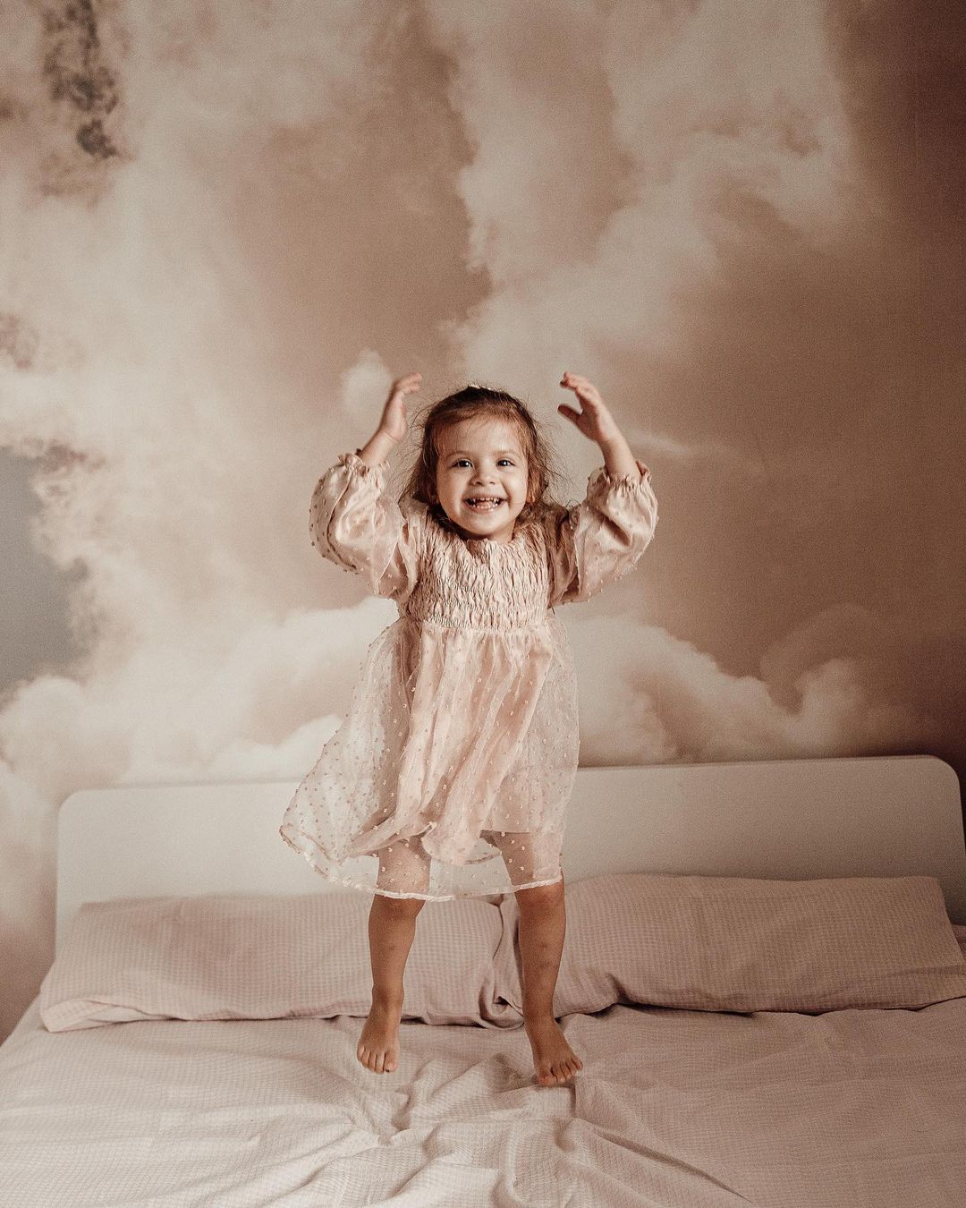 A smiling child in a pink dress jumps on a bed with a cloud-patterned wall behind.